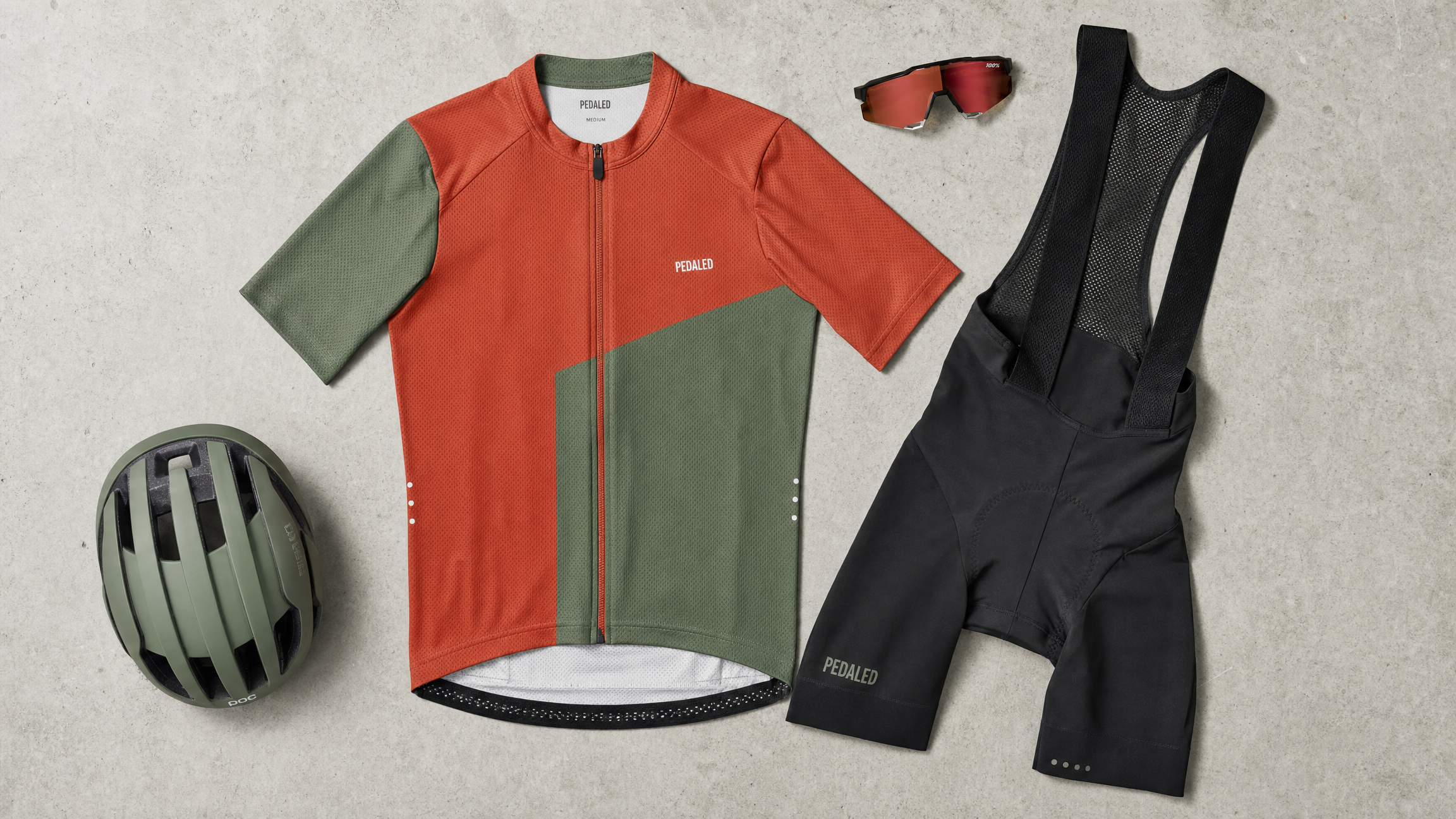 Flat lay of modern cycling apparel including a bold terracotta and sage green cycling jersey, black bib shorts, cycling sunglasses, and helmet on a light concrete surface, product photography style